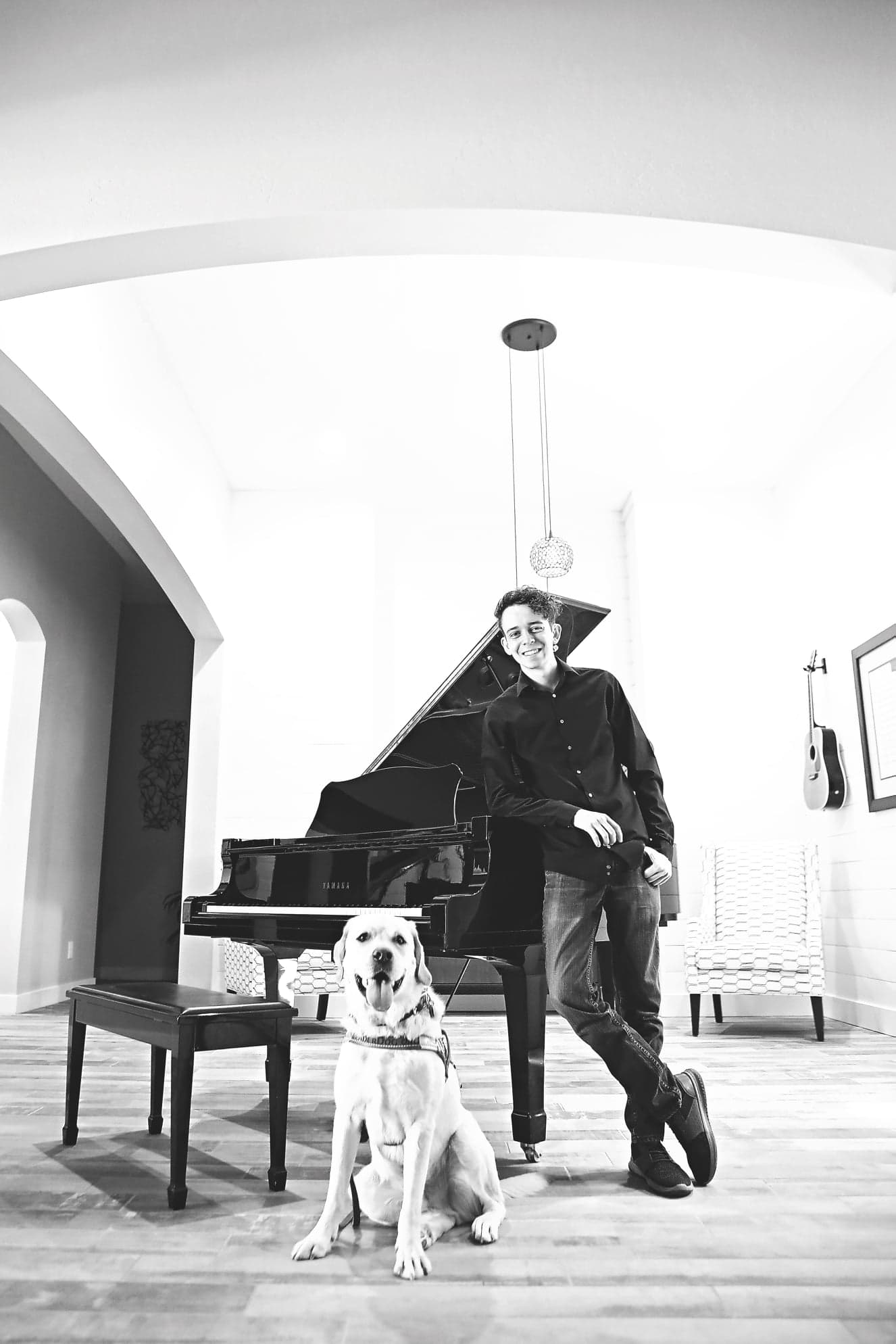 Brenden stands smiling, leaning against a black grand piano, with his guide dog Diesel sitting in front of him. Black-and-white photo.
