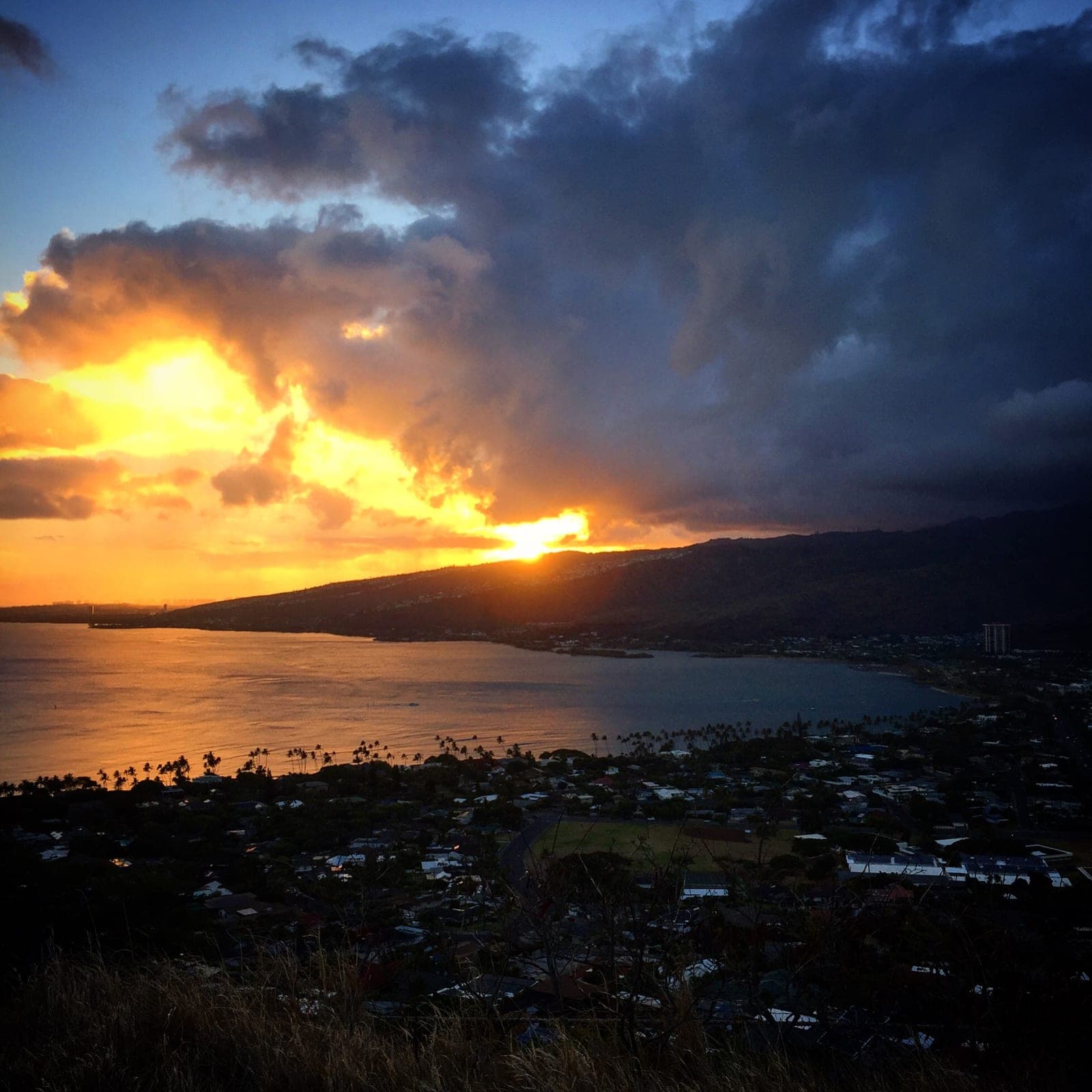 The sun breaks through dark storm clouds over a bay, lighting the water gold above a hillside town.