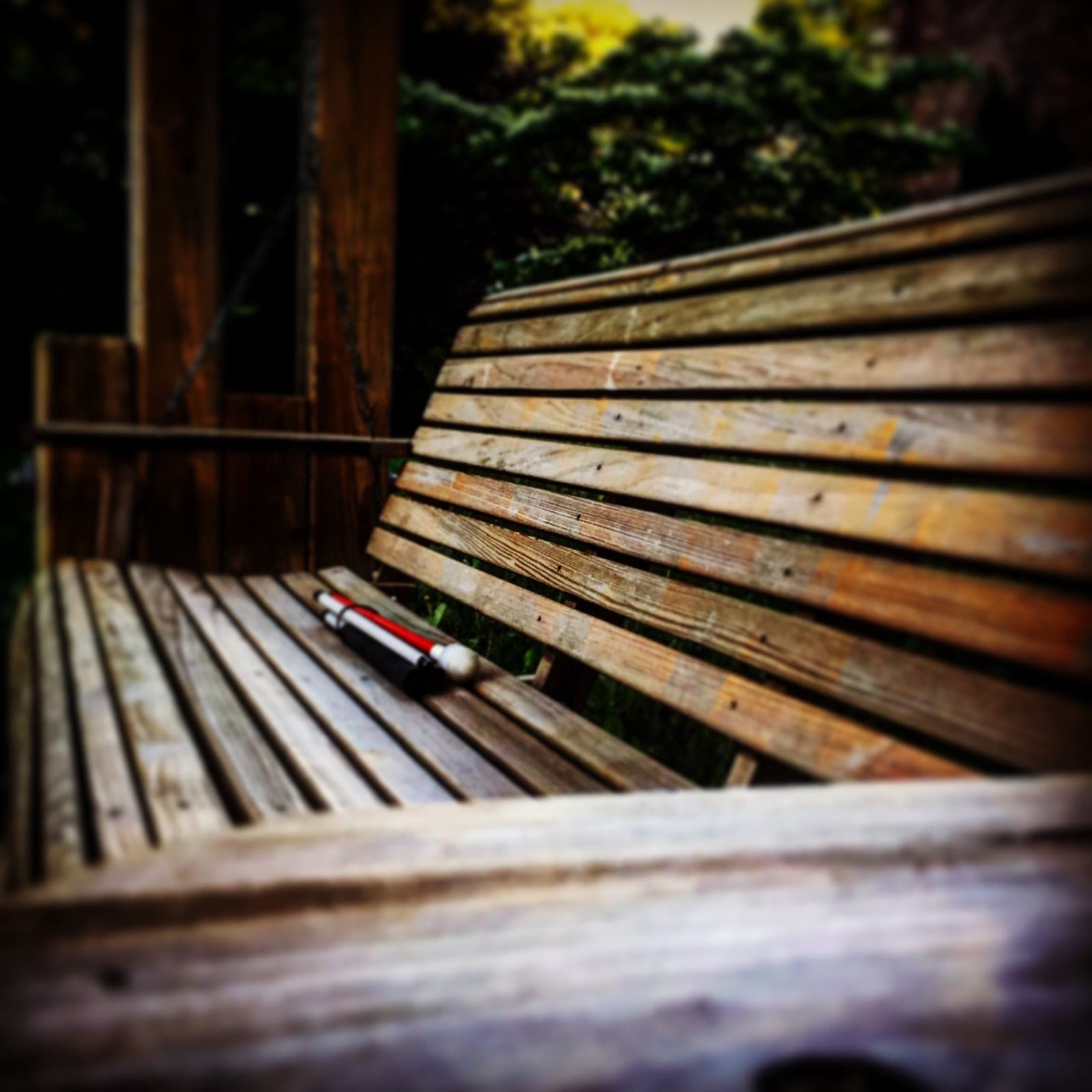 A white cane with a red stripe rests on the seat of a weathered wooden porch swing.